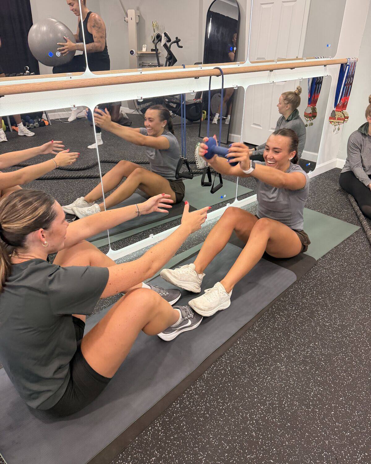 A small group on mats in front of a mirror, balancing in a V-sit and holding light weights